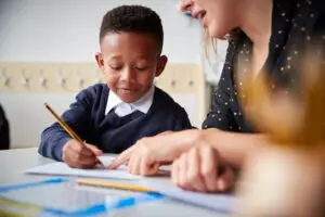 female primary school teacher helping a young school boy sitting at table in a classroom, close up, selective focus
