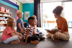 group of elementary school pupils sitting on floor listening to female teacher read story